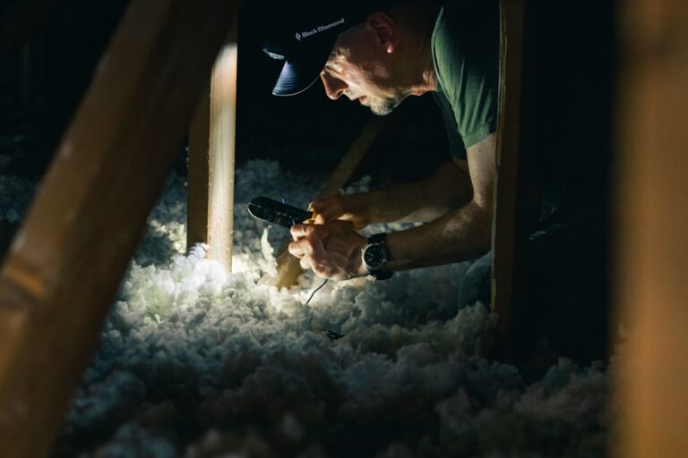 A homeowner in work clothes inspecting pink fiberglass insulation in a dimly lit residential attic.